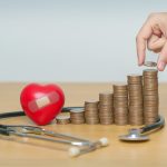 A hand putting money on a stack surrounded by a stethoscope
